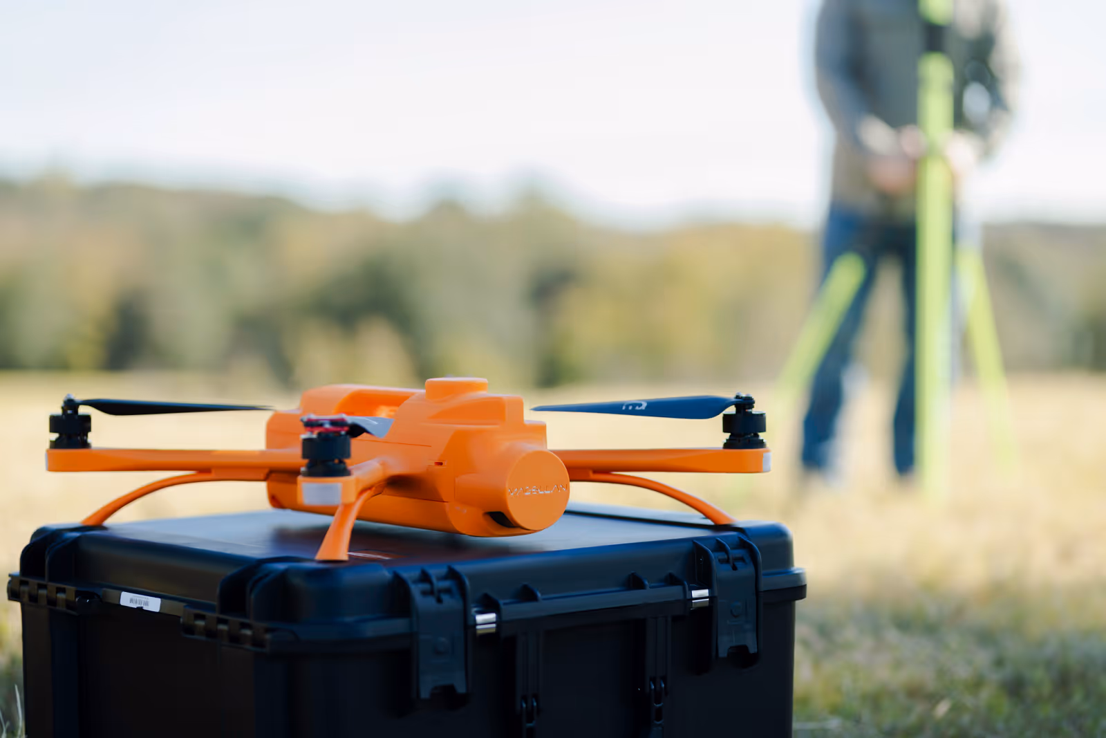 Orange drone resting on a black case outdoors with a person using surveying equipment blurred in background.