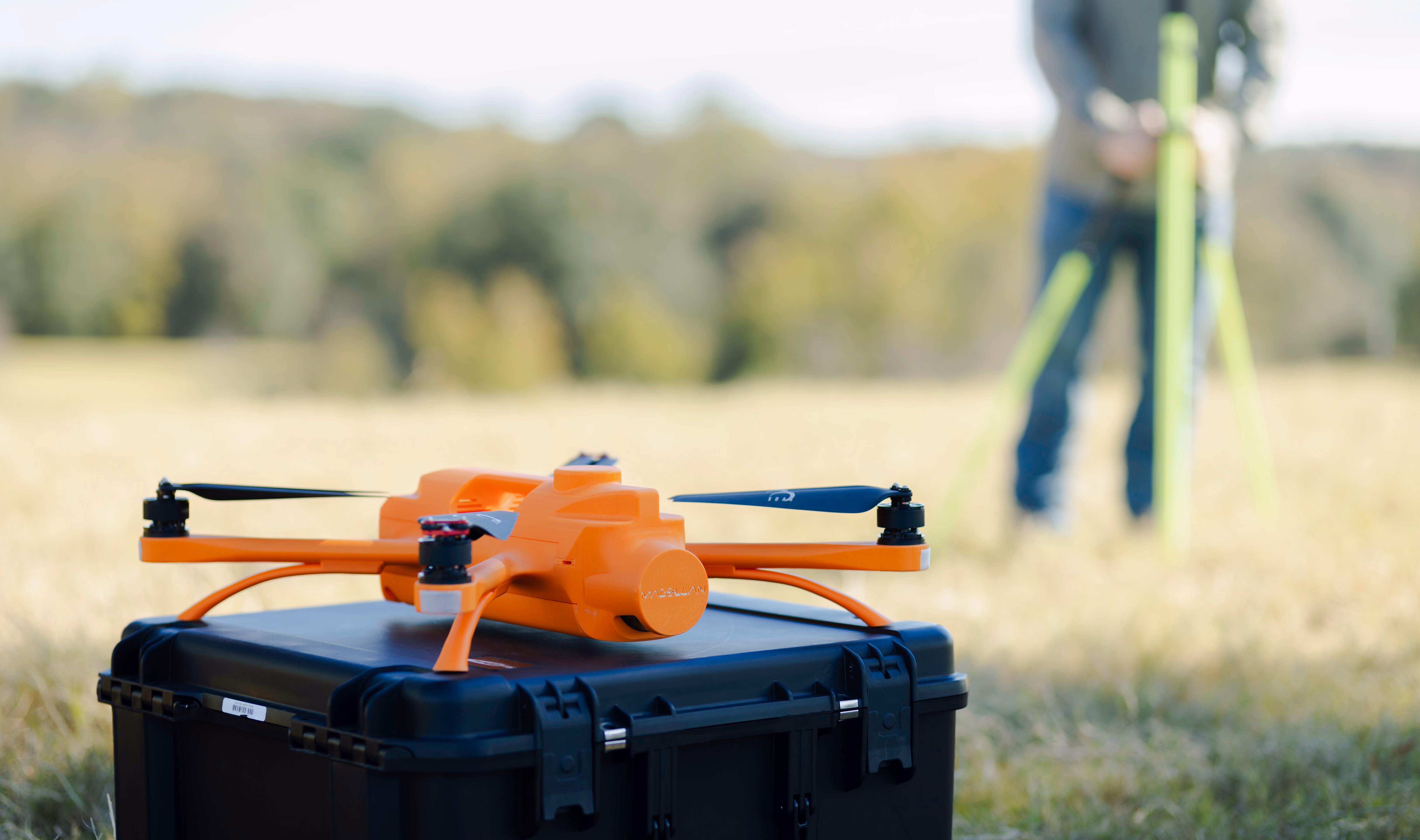 A SmartDrone Magellan surveying drone sits on its case while a land surveyor sets up a base station in the background