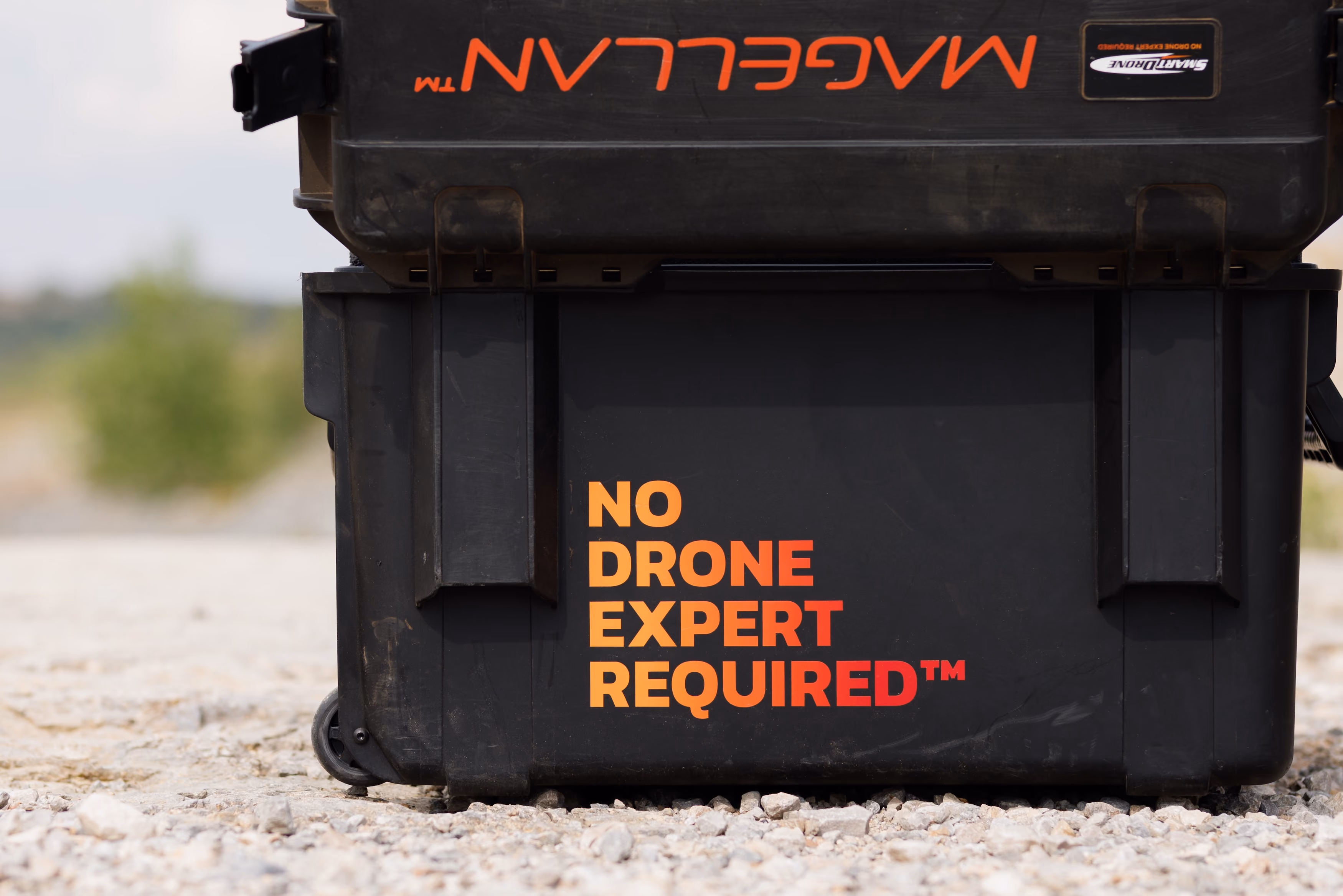 Close-up of black rugged cases stacked outdoors on rocky ground, with bold orange and red text on the bottom case stating 'NO DRONE EXPERT REQUIRED™' and 'MAGELLAN' on the top case.