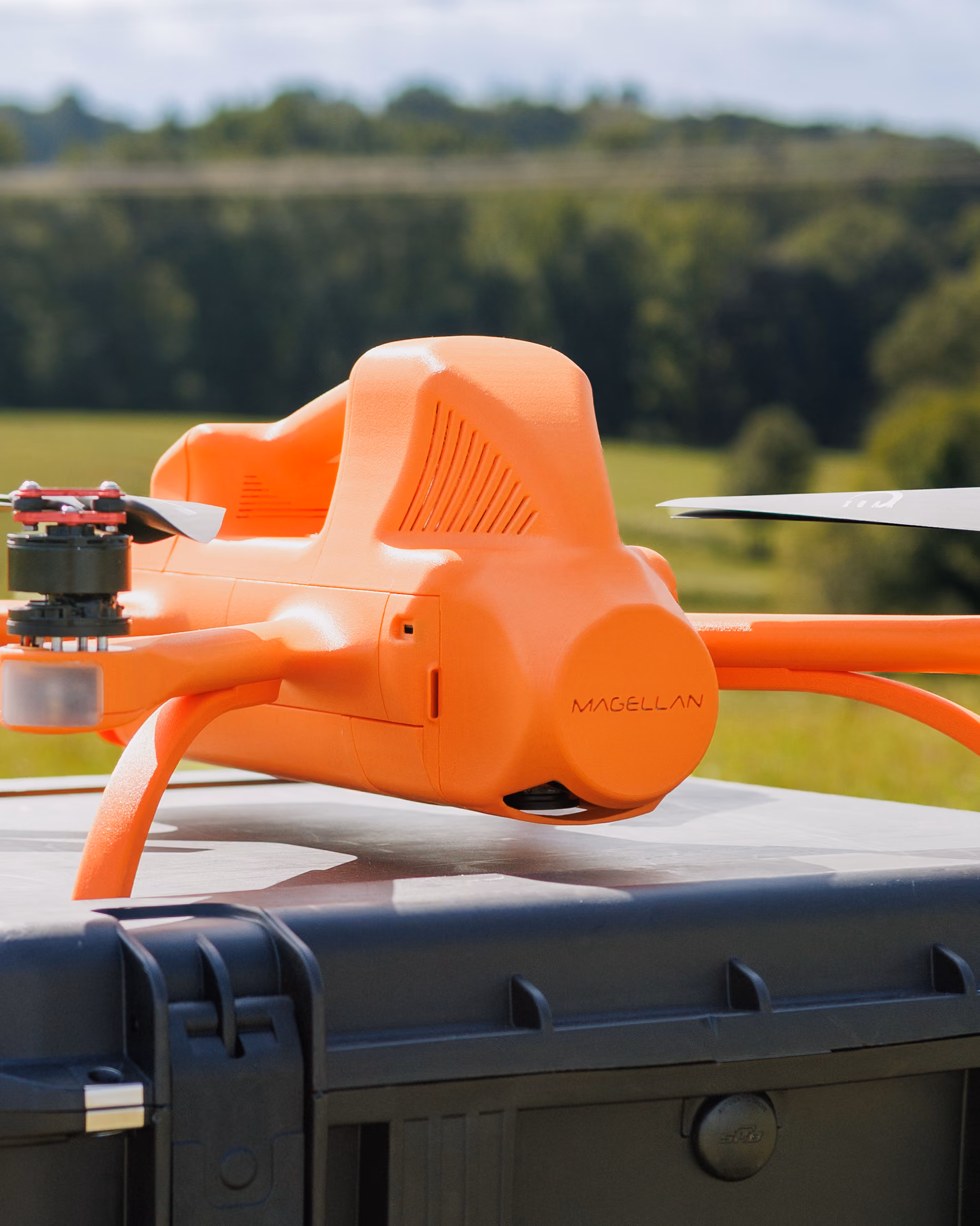 Close-up of an orange Magellan drone resting on a black case outdoors with blurred green trees in the background.