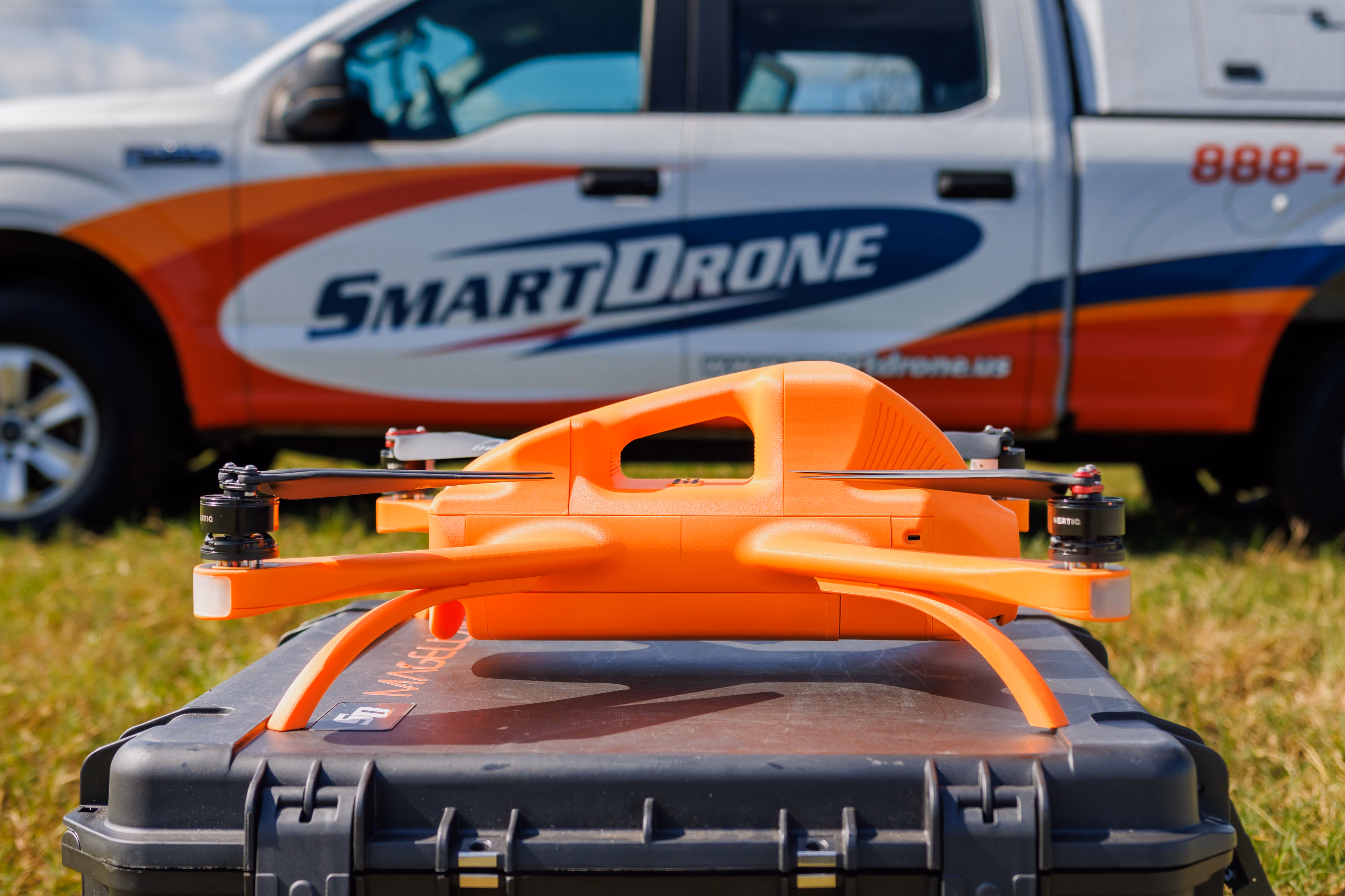 Man in casual attire carrying an orange drone walking beside a white SmartDrone truck in a grassy field.
