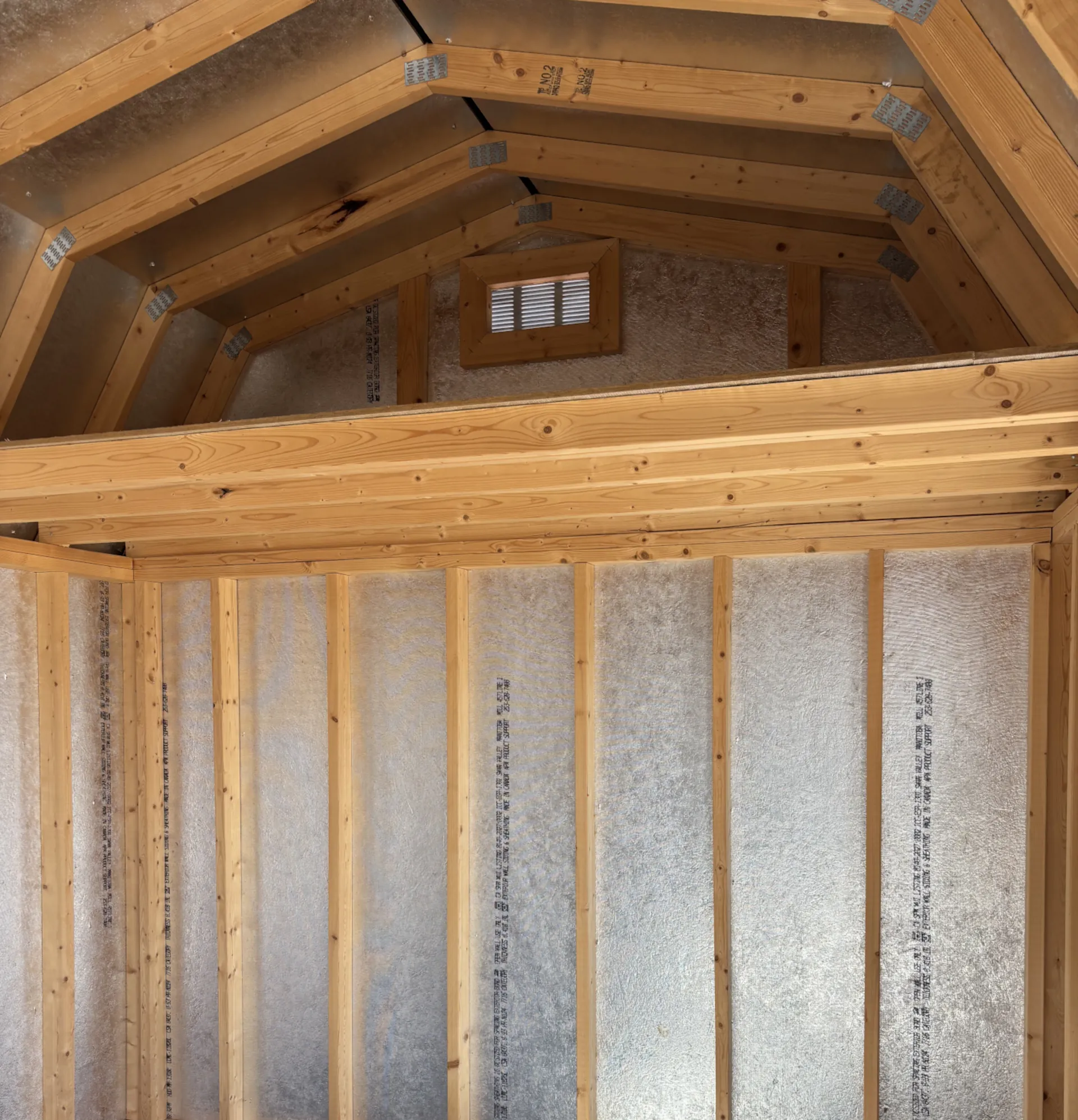 Interior view of a wooden gambrel roof shed with loft framing and a small vented window.