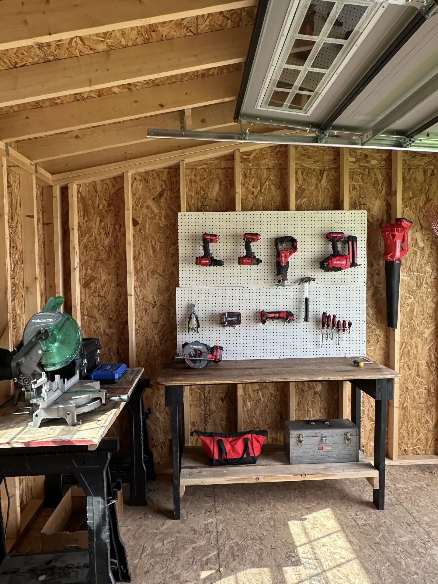 Workbench in wooden shed with power tools on pegboard and saw on nearby table.