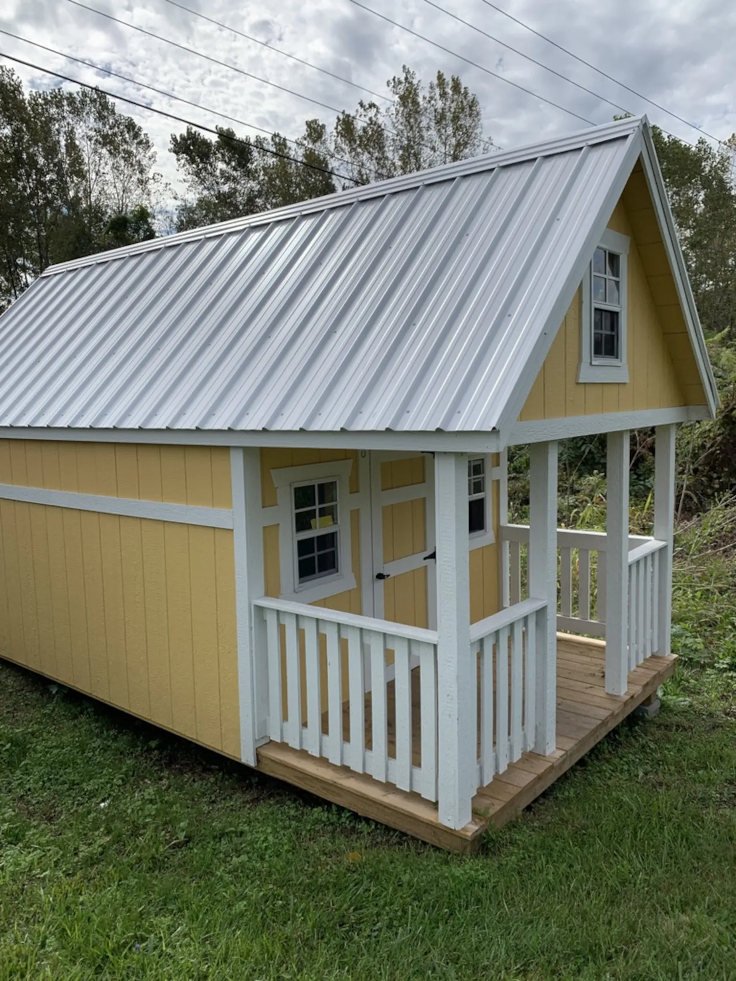 Yellow wooden playhouse shed with white trim, metal roof, small porch, and windows on grassy ground.
