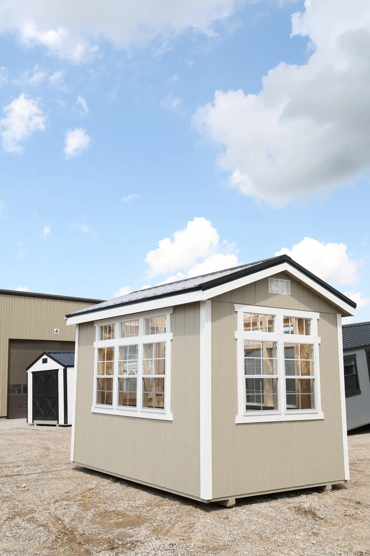 Beige greenhouse shed with white-trimmed windows on a gravel lot under a partly cloudy blue sky.