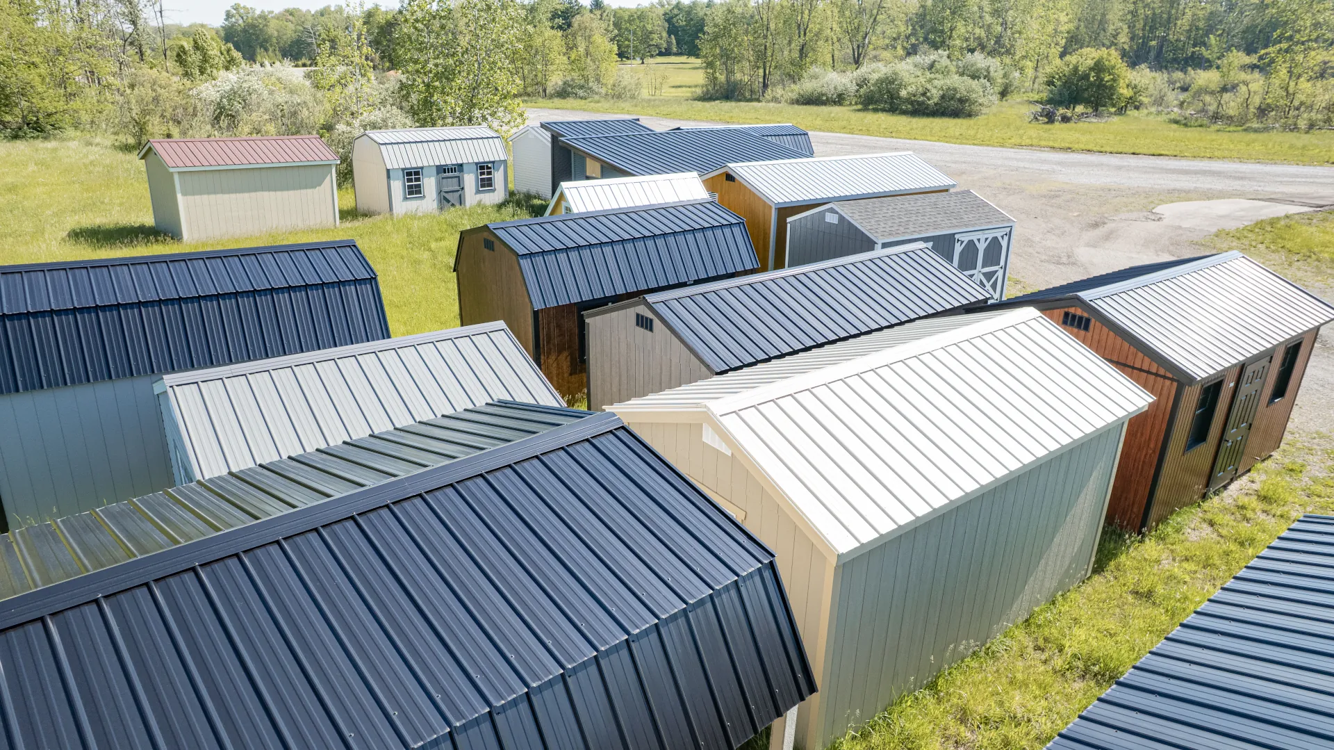 Multiple outdoor sheds with metal roofs on green grass with trees and a gravel road in background.