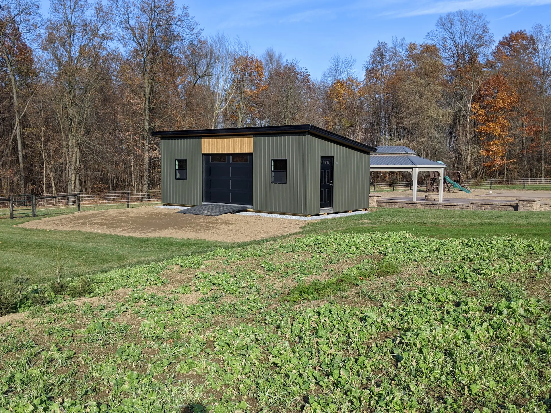Green metal shed with black garage door and ramp, standing on soil with trees and a playground nearby.