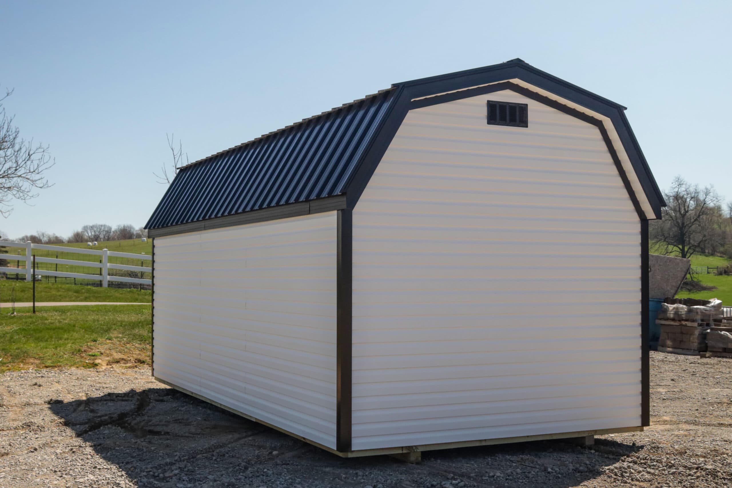 White and black metal-roofed shed on gravel with grassy field and fence in background.