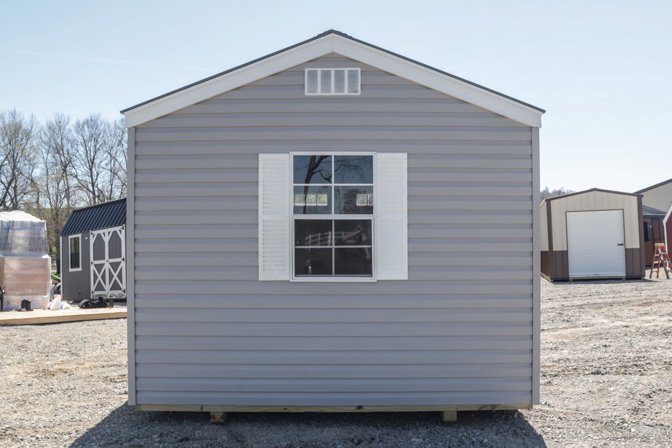 Gray siding building with a white window and shutters on gravel under clear sky.