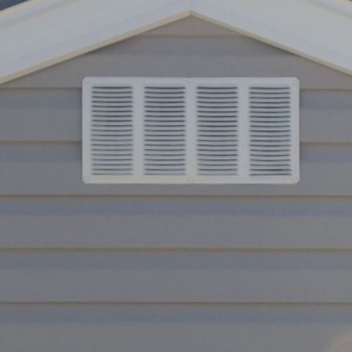 White rectangular vent with horizontal slats on gray siding below a gable roof.