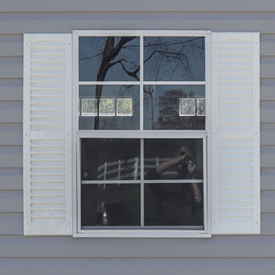 White framed window with closed shutters reflecting trees and a person taking a photo.