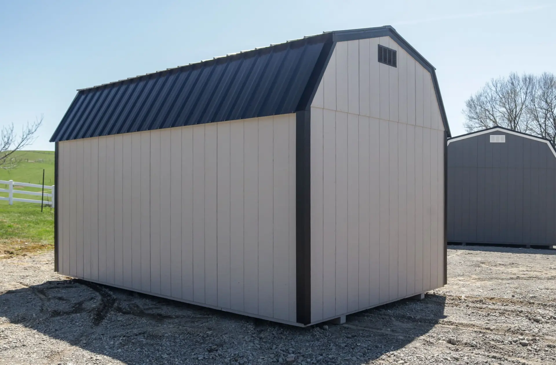 Beige storage shed with a black metal gambrel roof sits on gravel with another gray shed in the background.