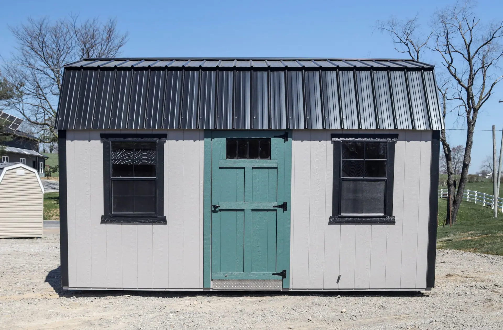 Small beige shed with teal door, black roof and window frames on a gravel surface outdoors.