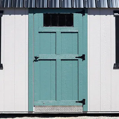 Teal single wooden door with black hinges set in white building with corrugated metal roof.