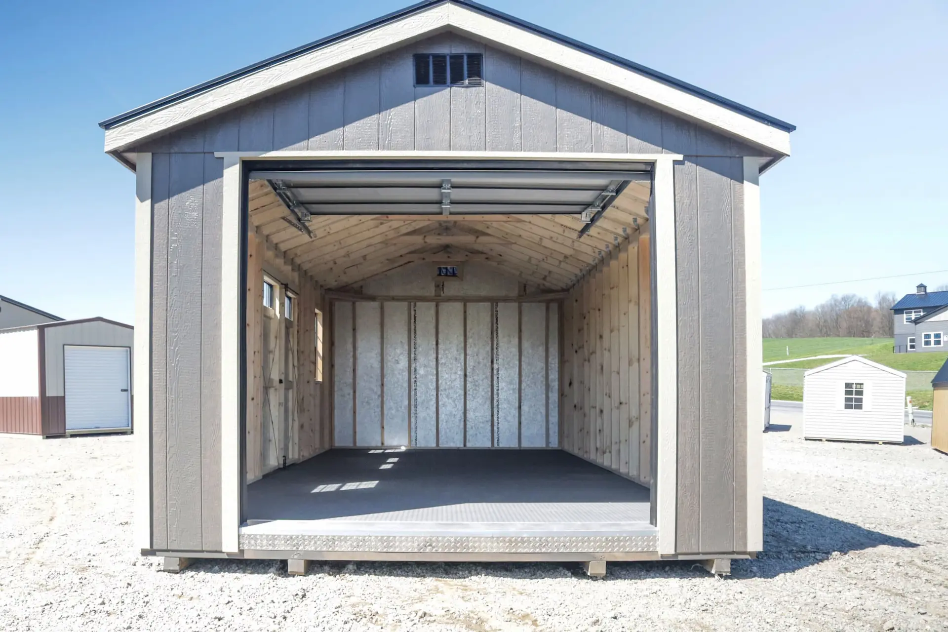 Empty gray wooden storage shed with open garage-style door on a gravel lot.