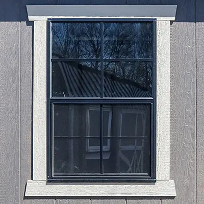 A window with white trim on gray siding reflecting trees and a roof in the glass panels.