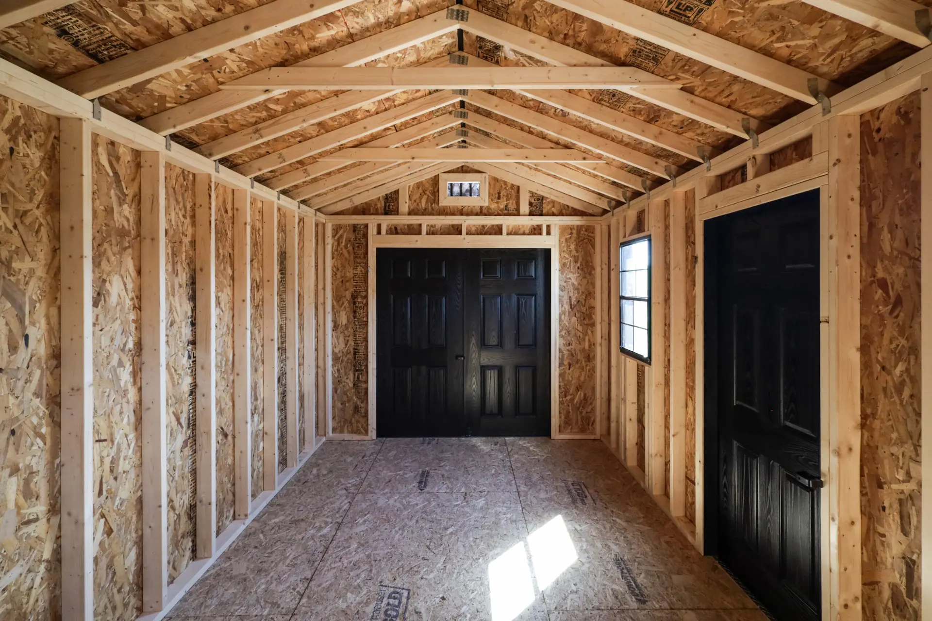 Empty wooden shed interior with exposed framing, black double doors at the end, a window, and one black door on the side.