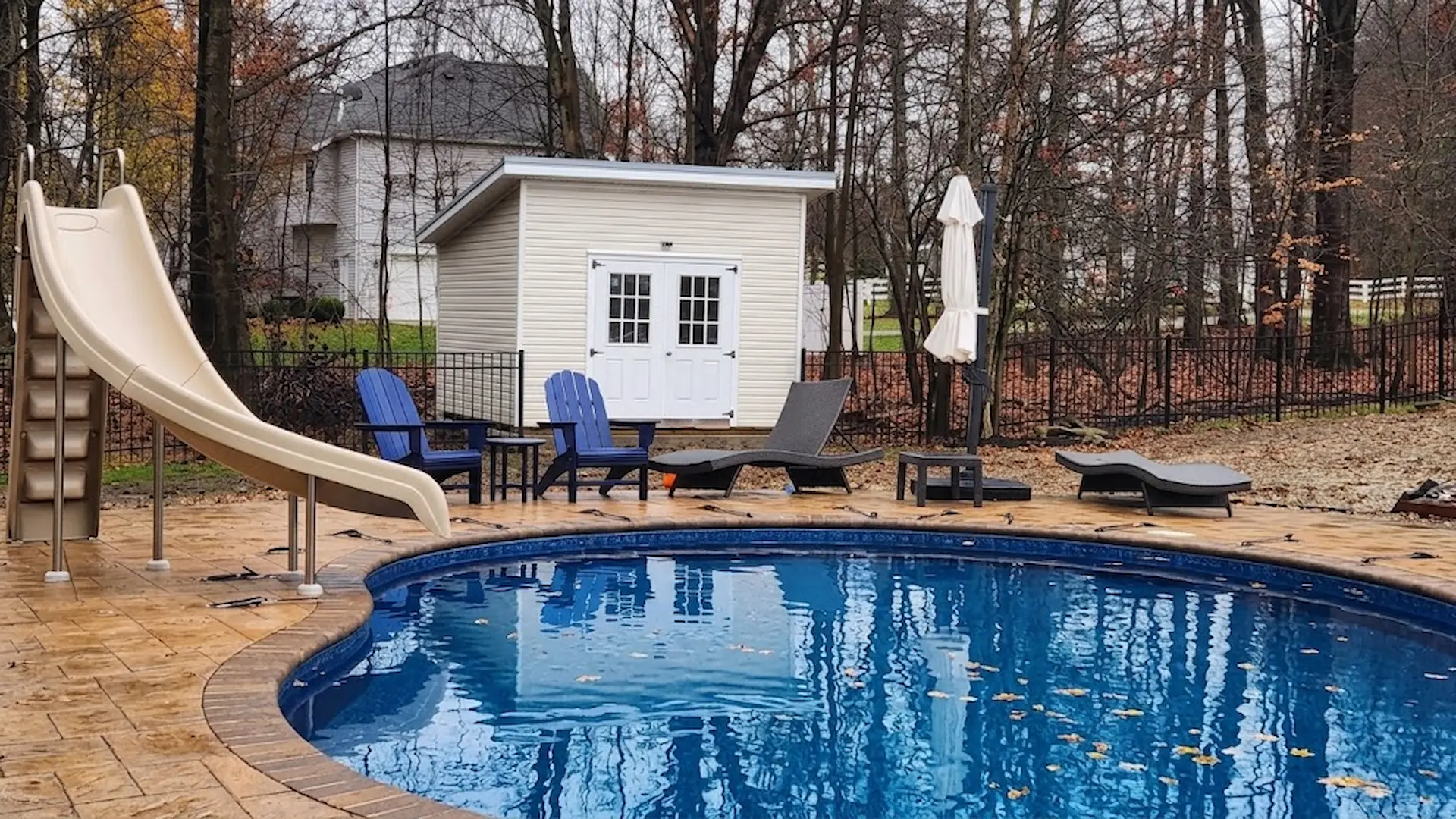 Outdoor scene of a backyard pool area with a beige slide leading into a blue swimming pool, two blue Adirondack chairs, a gray lounge chair, an umbrella, and a beige storage shed in the background surrounded by leafless trees and a black metal fence.