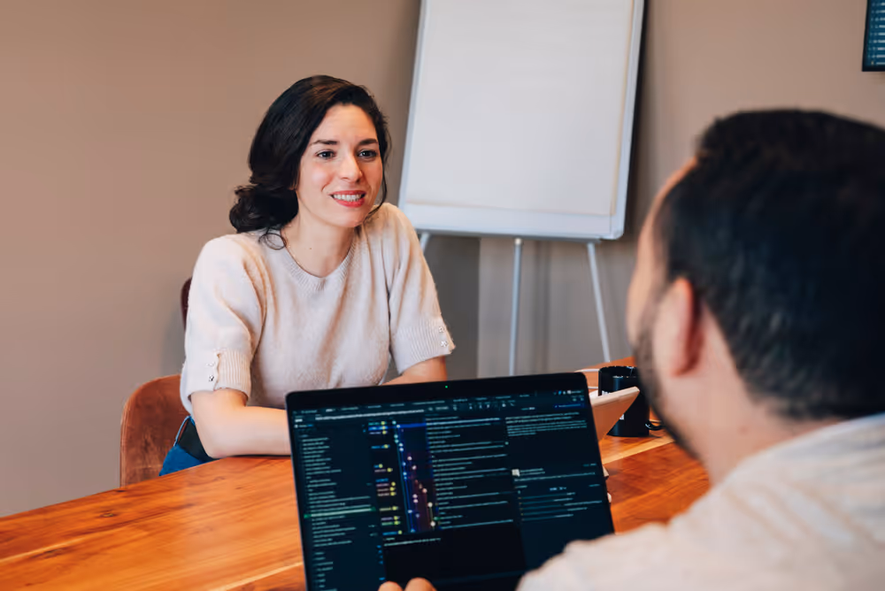 Woman smiling and talking to a man working on a laptop in an office room with a whiteboard.