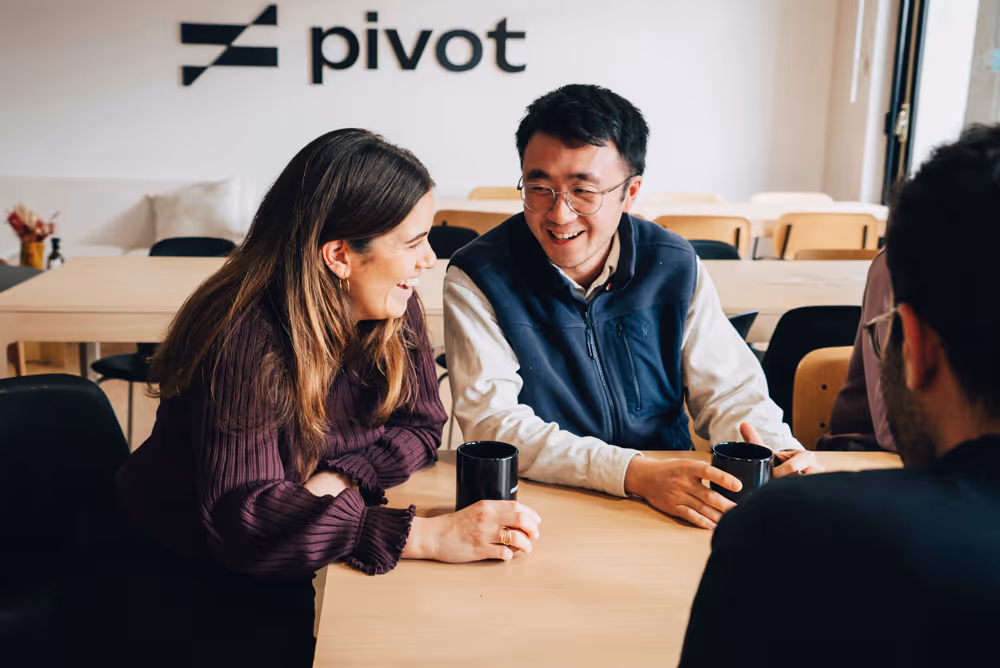 Three colleagues smiling and talking while holding black mugs at a wooden table in an office with Pivot logo.