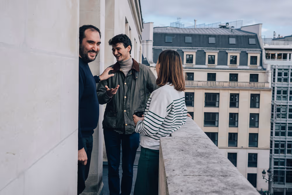 Three people talking and laughing on a balcony overlooking city buildings on a cloudy day.