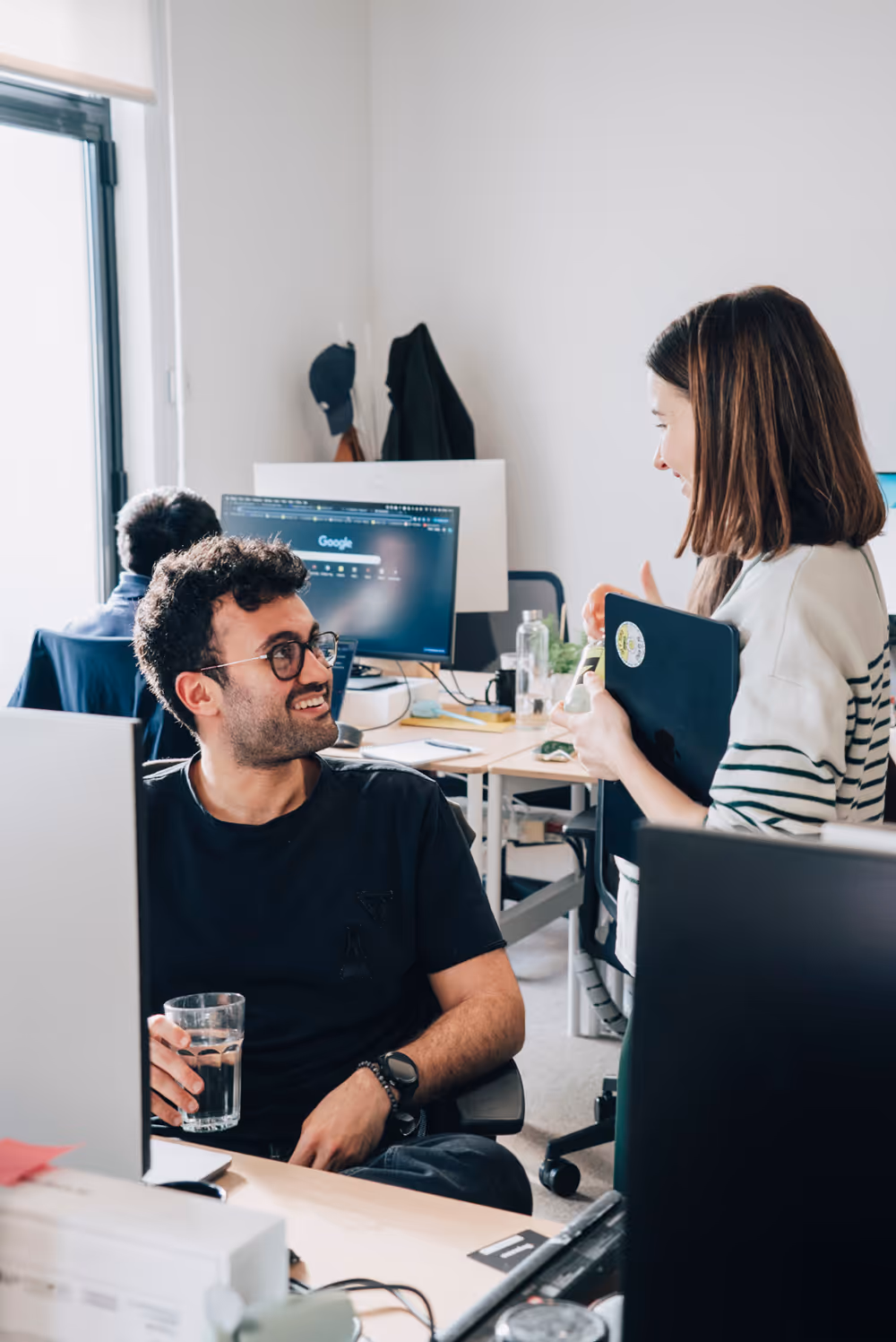 Man sitting at desk holding a glass of water, smiling at a woman standing with a laptop in an office.