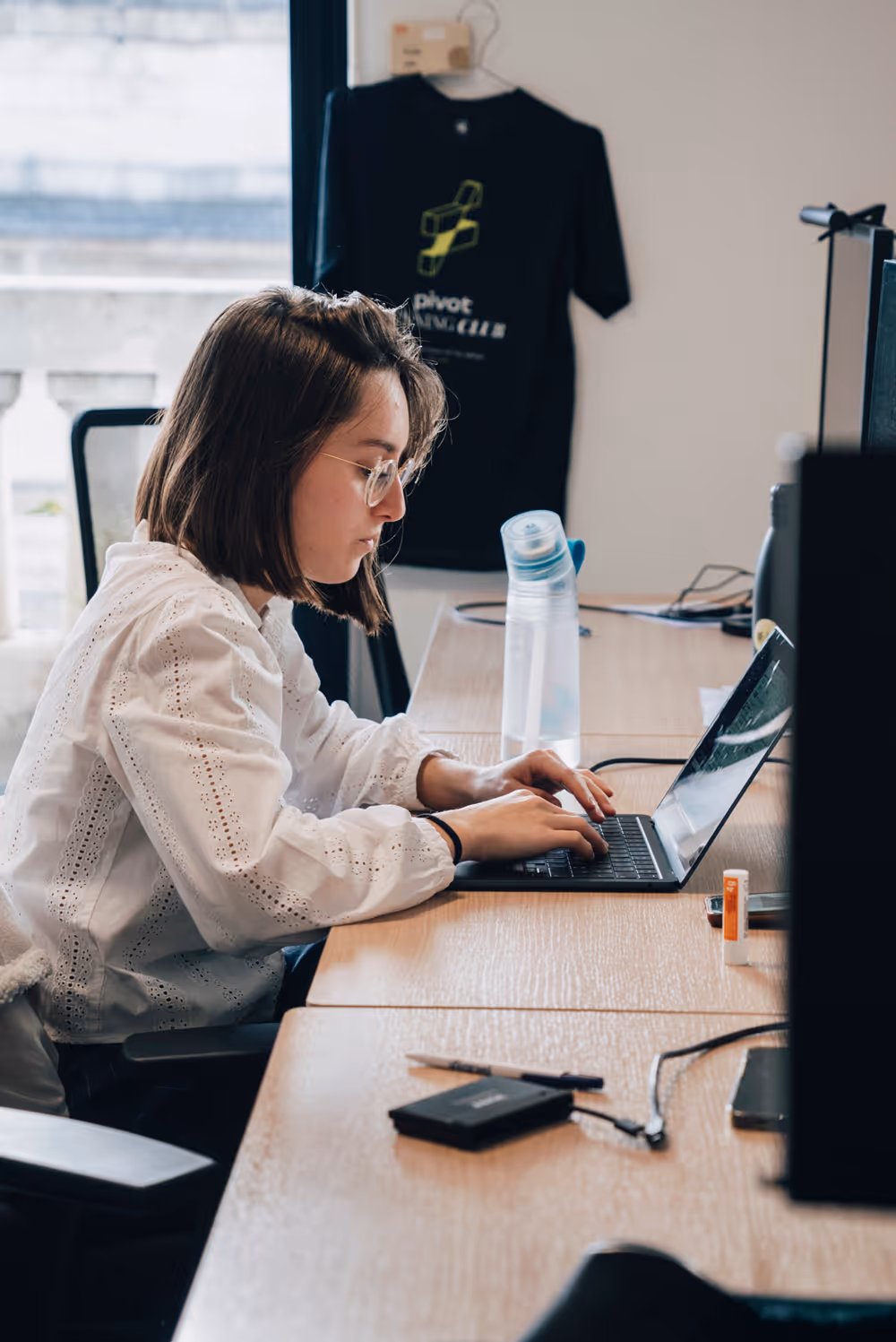 Young woman wearing glasses typing on a laptop at a wooden desk in an office.