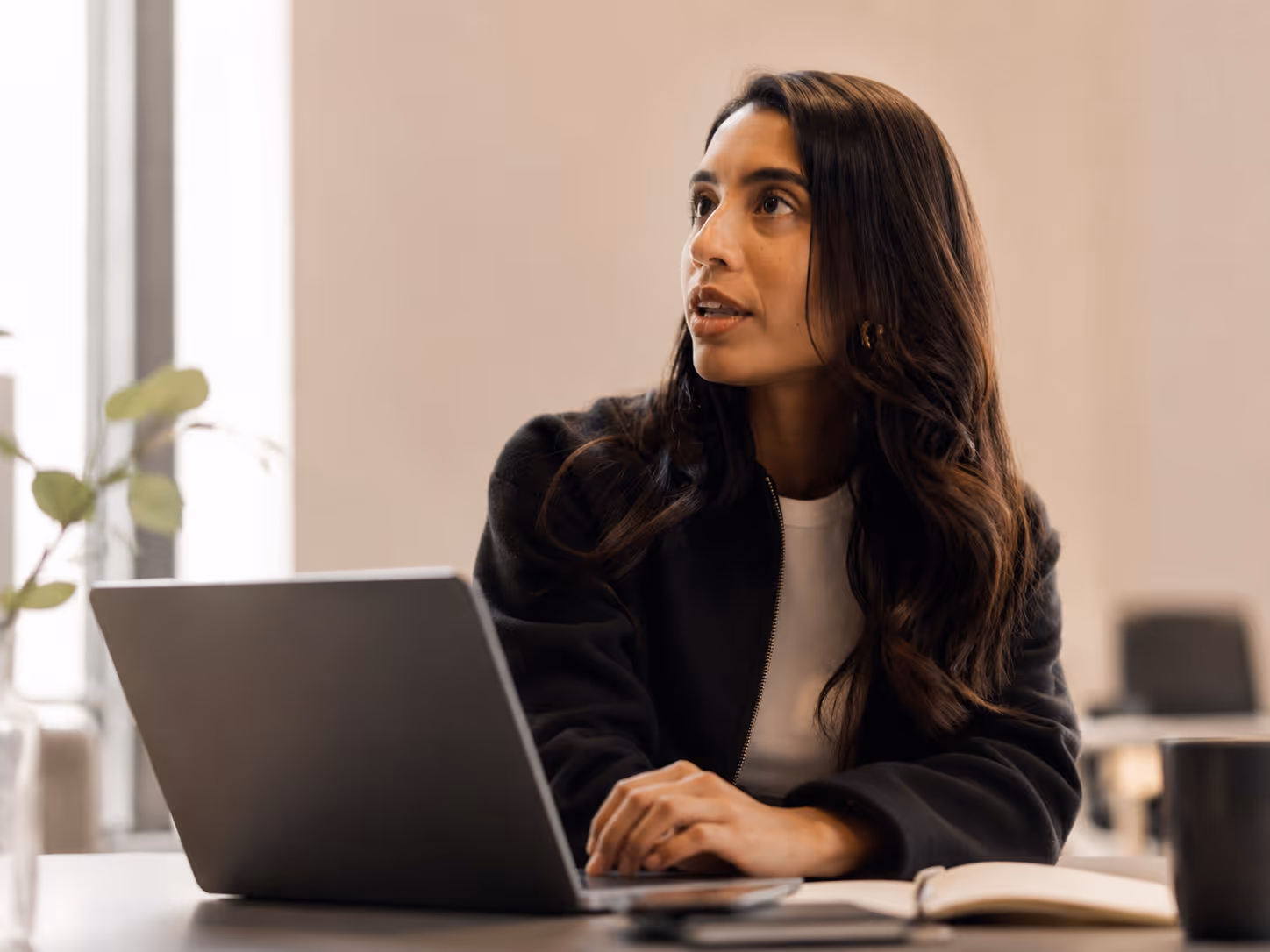 Woman with long dark hair sitting at a table using a laptop, looking attentively to the side.