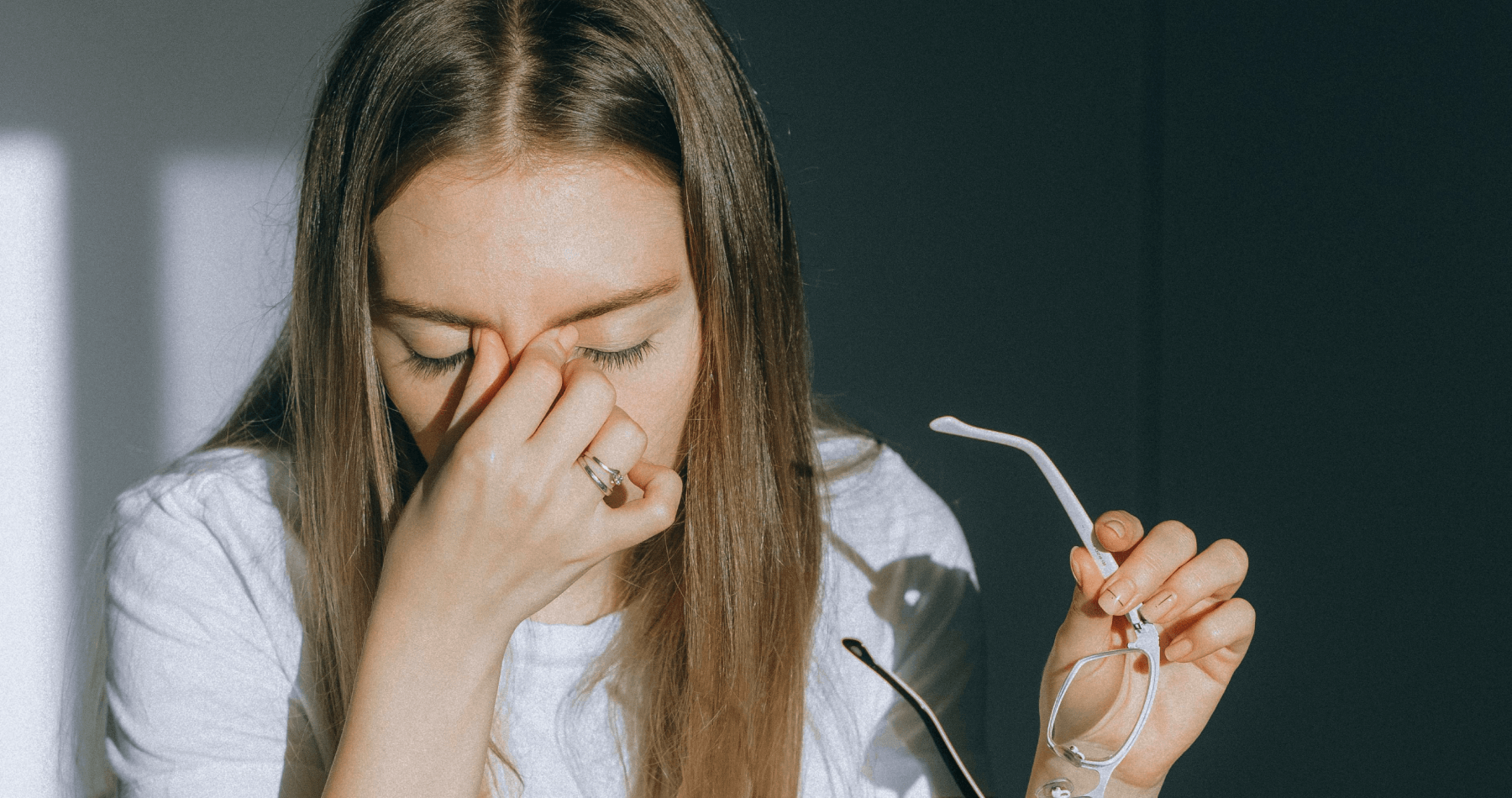 A woman closes her eyes and pinches the bridge of her nose while holding her glasses, showing signs of eye strain or fatigue.