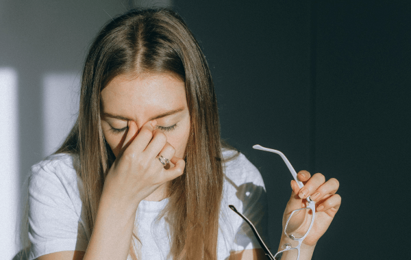 A woman closes her eyes and pinches the bridge of her nose while holding her glasses, showing signs of eye strain or fatigue.