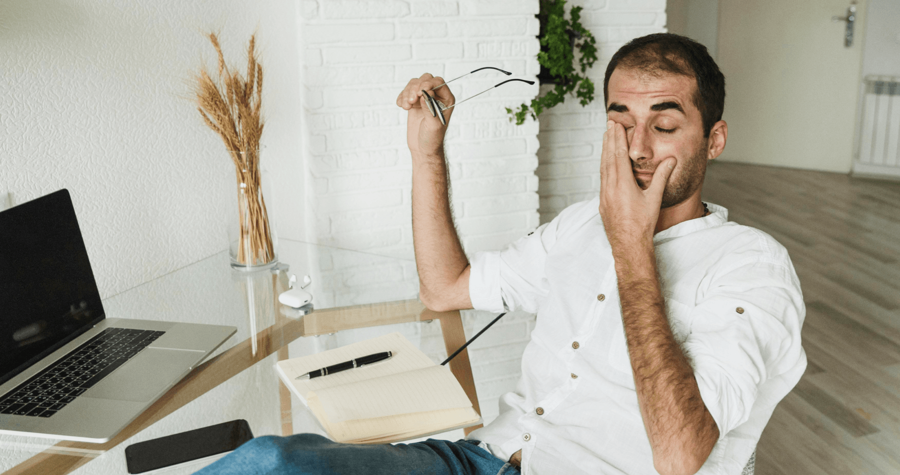 Man sitting at a desk rubbing his eye while holding glasses, showing signs of eye strain from working on a laptop.