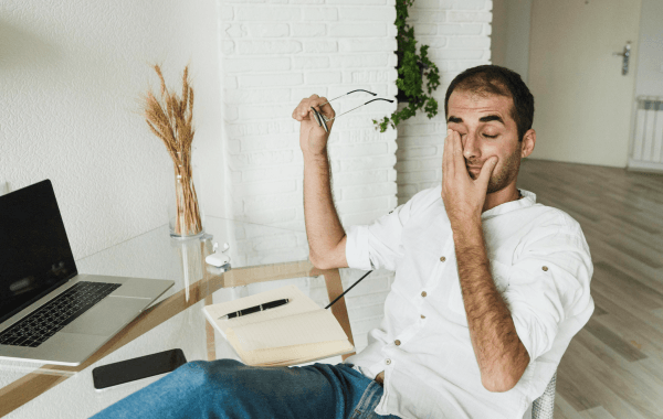 Man sitting at a desk rubbing his eye while holding glasses, showing signs of eye strain from working on a laptop.