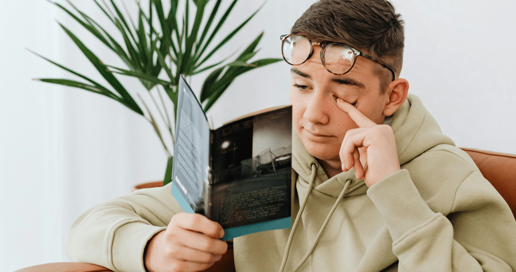 Teen wearing glasses and a hoodie rubbing one eye while reading a book indoors, illustrating eye strain and discomfort from prolonged reading.