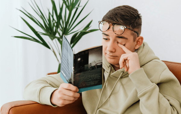 Teen wearing glasses and a hoodie rubbing one eye while reading a book indoors, illustrating eye strain and discomfort from prolonged reading.