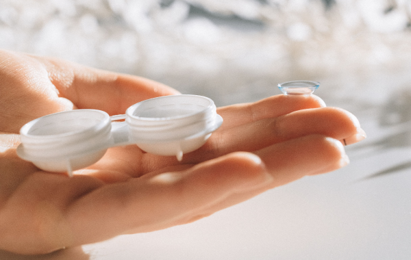 A clear soft contact lens balancing on a person's index fingertip next to an open white contact lens storage case.