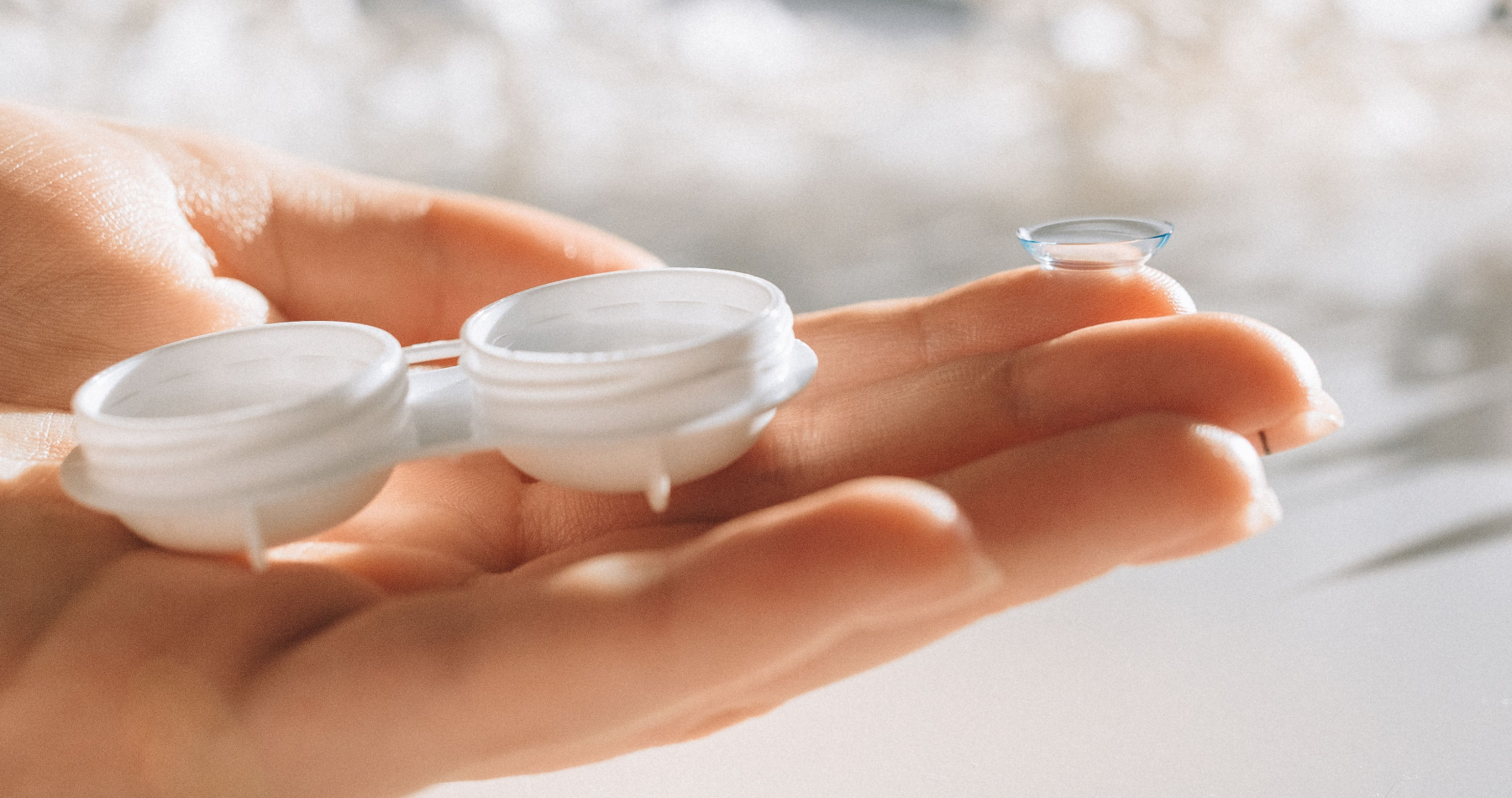 A clear soft contact lens balancing on a person's index fingertip next to an open white contact lens storage case.