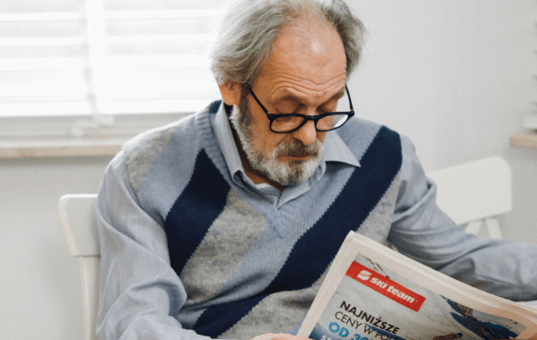 Senior man wearing eyeglasses holding and looking down at a newspaper, illustrating age-related vision changes or presbyopia.