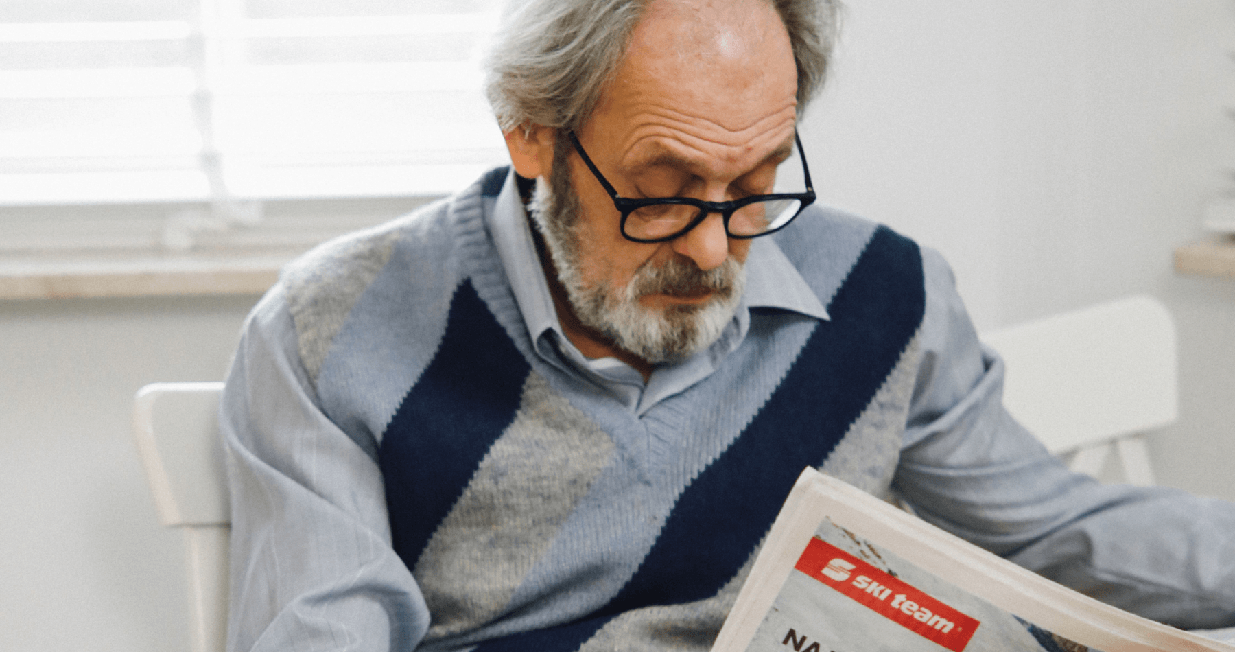Senior man wearing eyeglasses holding and looking down at a newspaper, illustrating age-related vision changes or presbyopia.