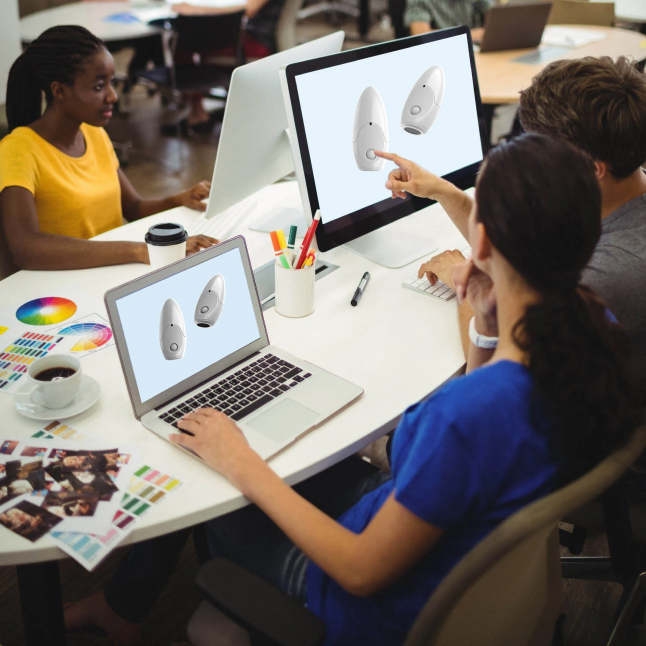 In a collaborative workspace within a light therapy company, several people are actively involved in product - related activities. A woman in a blue top is pointing at a computer monitor displaying images of a white - colored device, likely related to light therapy products. Another individual is working on a laptop that also shows the same device. The table is cluttered with color palettes, coffee cups, and stationery, suggesting a creative environment focused on the design and development of light therapy products. 