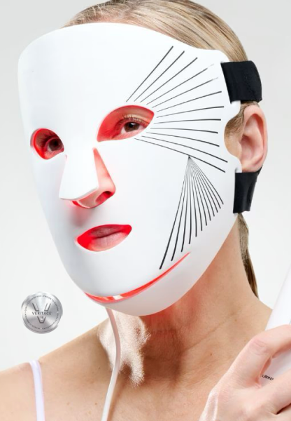 A woman wearing a white lablel facial mask with red LED light accents and black strap, featuring black line patterns radiating from the eyes, holding a power cord, with a 'Veritace®' silver badge in the lower left corner