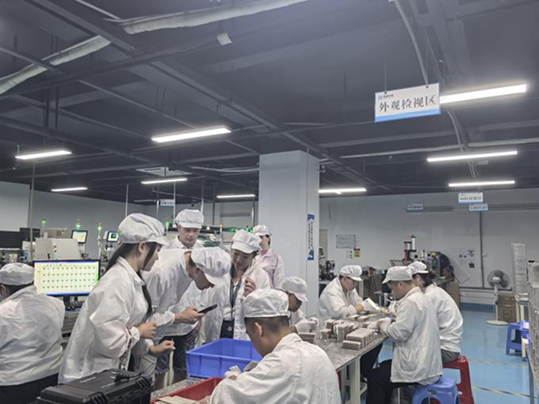 Factory interior of a light therapy manufacture plant, showing workers in white attire gathered at the 'Appearance Inspection Area' for production - related tasks.