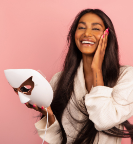 A woman in a white robe holds a white LED facial mask, against a pink background, with one hand touching her cheek