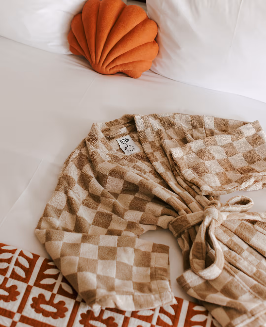 A beige checkered robe lies on a bed with white sheets, an orange pillow, and a patterned blanket.