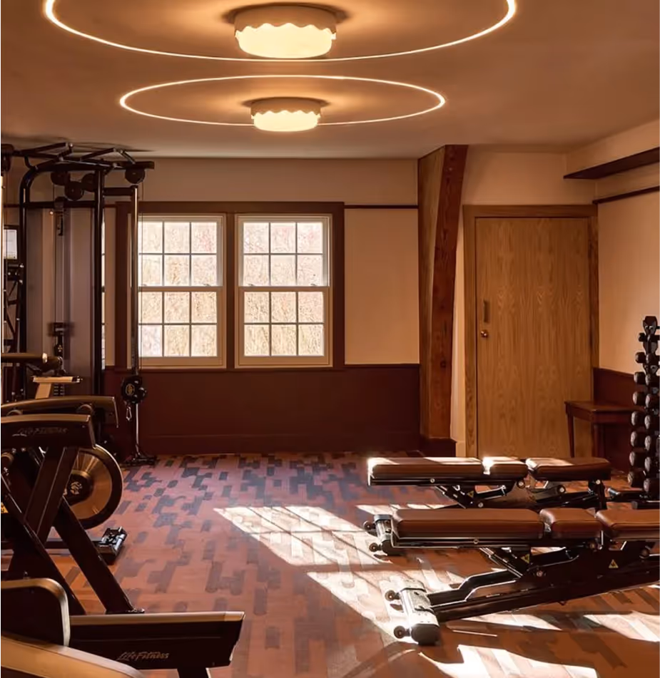 A gym room with workout machines, benches, dumbbells, and large windows letting in natural light.