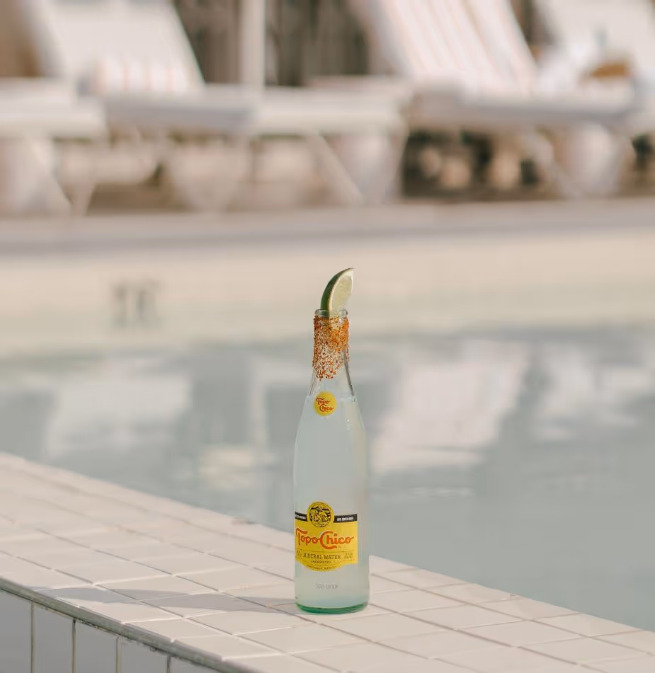 A Topo Chico bottle with a lime wedge and chili powder on the rim sits on the edge of a pool with lounge chairs in the background.