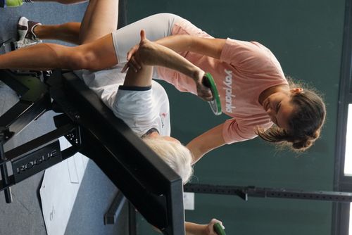 A woman smiles and assists a man who is lying on the floor in a gym, raising his arms as if exercising. She is wearing a peach-colored shirt with "yay planks" text. Gym equipment is visible in the background. Both look engaged and focused, showcasing personal training in Palma Illes Balears.