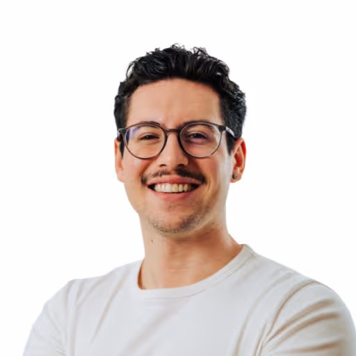 Lächelnder Mann mit kurzen schwarzen Locken, Brille und weißem T-Shirt vor weißem Hintergrund.