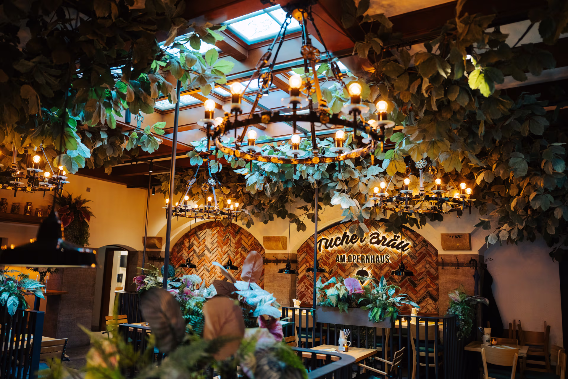 Gemütliches Restaurant mit warmem Licht, großen Kronleuchtern und üppigen grünen Pflanzen an der Decke und auf Tischen.