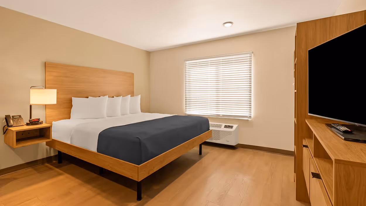A hotel room with a king bed, wooden furniture, a TV, nightstand, and window with closed blinds.