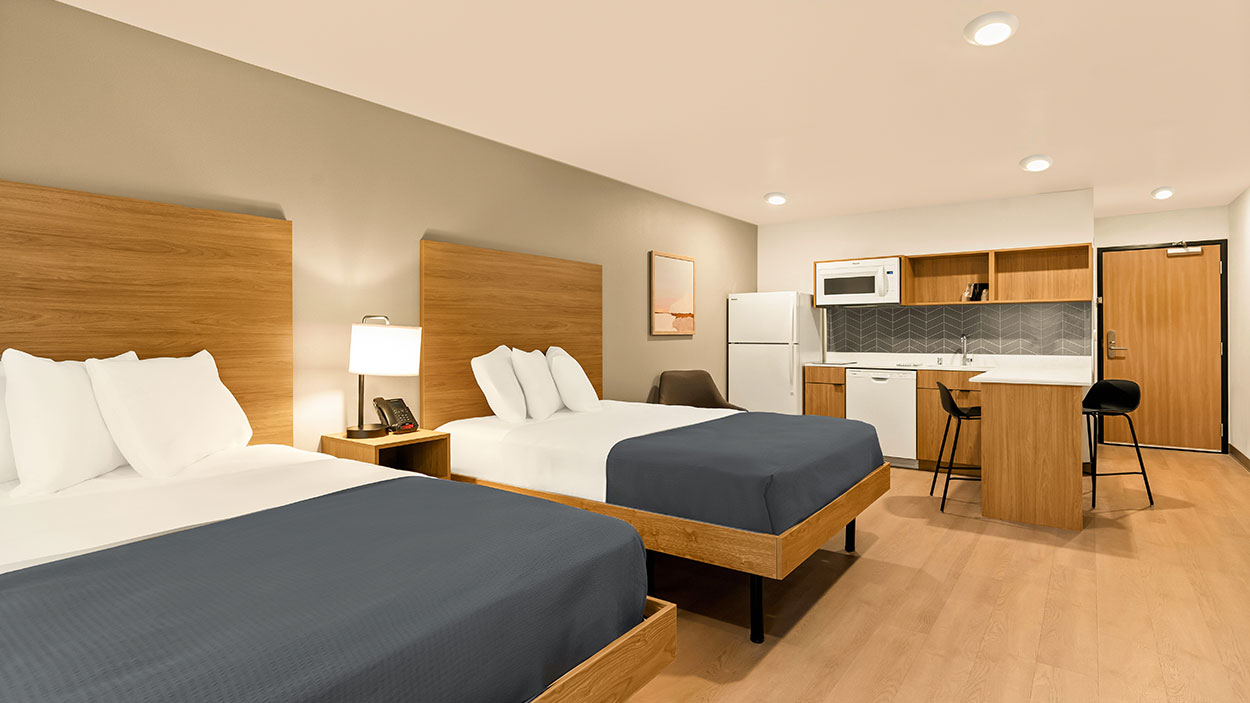 Hotel room with two double beds, a kitchenette, dining area, and wood flooring under bright ceiling lights.